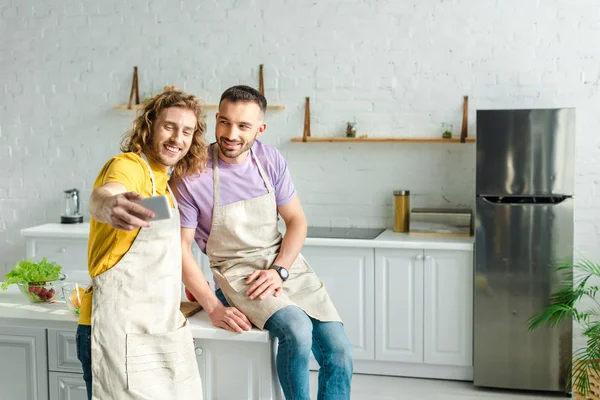 Foco seletivo de homens homossexuais felizes em aventais tomando selfie na cozinha — Fotografia de Stock