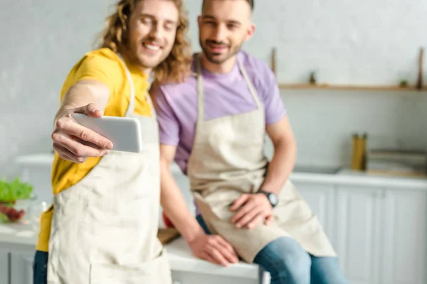 Selective focus of cheerful homosexual men in aprons taking selfie — Stock Photo