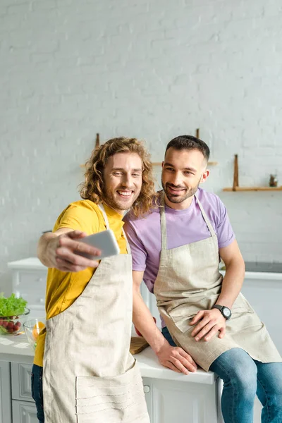 Enfoque selectivo de hombres homosexuales felices en delantales tomando selfie - foto de stock