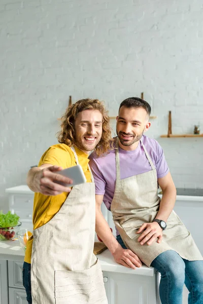 Enfoque selectivo de hombres homosexuales felices en delantales tomando selfie - foto de stock