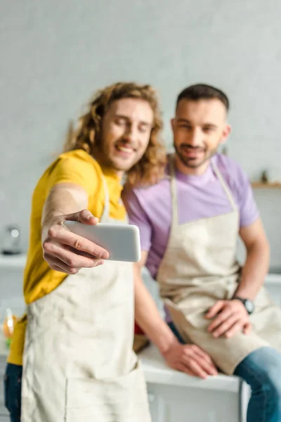 Selective focus of homosexual men in aprons taking selfie — Stock Photo