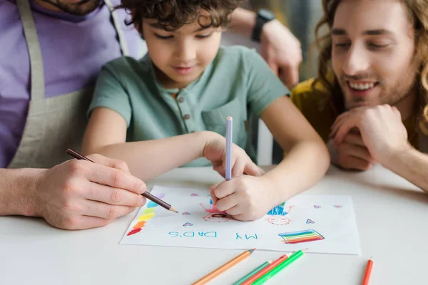 Mischlingskind zeichnet Bild mit Papas Schriftzug in der Nähe glücklicher homosexueller Eltern — Stockfoto