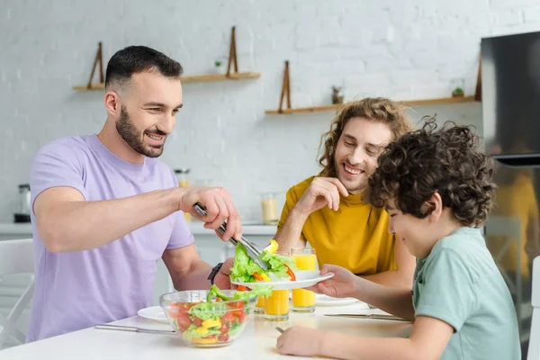 Feliz hombre homosexual poniendo ensalada en plato de raza mixta hijo - foto de stock