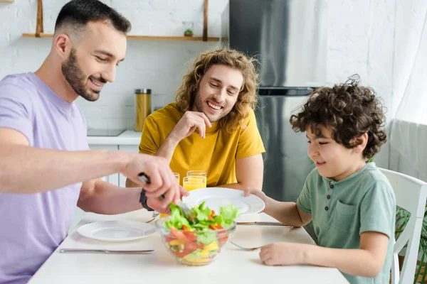 Hombres homosexuales felices y niño de raza mixta mirando sabrosa ensalada - foto de stock