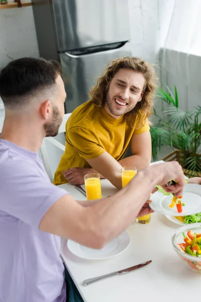Homosexual and bearded man putting salad on plate of happy husband — Stock Photo
