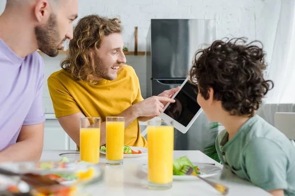 Hombre homosexual feliz apuntando con el dedo a la tableta digital con la pantalla en blanco cerca del hijo de raza mixta - foto de stock