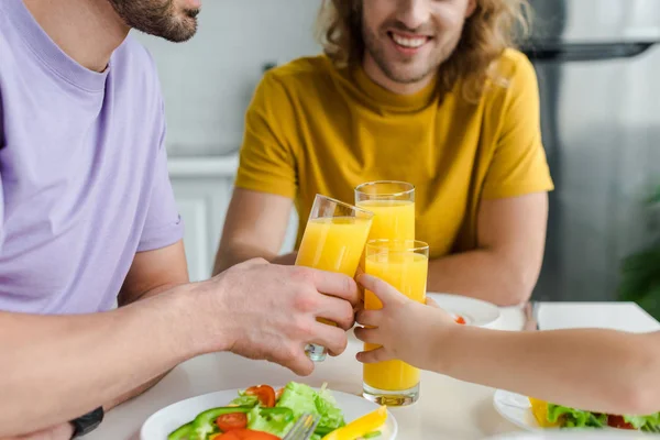 Vista recortada de los hombres homosexuales felices y el hijo tintineo vasos con jugo de naranja — Stock Photo