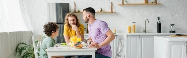 Plano panorámico de los padres homosexuales y lindo hijo de raza mixta almorzando en casa - foto de stock