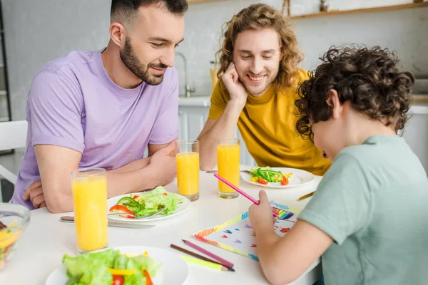 Mischlingskind zeichnet Bild mit lgbt-Fahne in der Nähe glücklicher homosexueller Eltern — Stockfoto