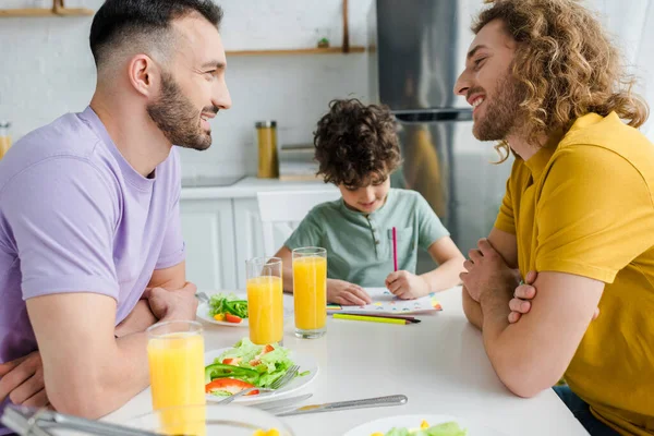 Selective focus of happy homosexual parents looking at each other near mixed race son drawing picture — Stock Photo