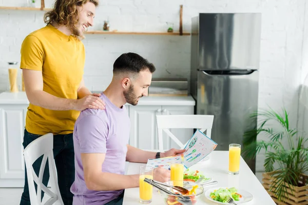 Homens homossexuais felizes olhando para papéis com desenho perto de salada e óculos com suco de laranja — Fotografia de Stock