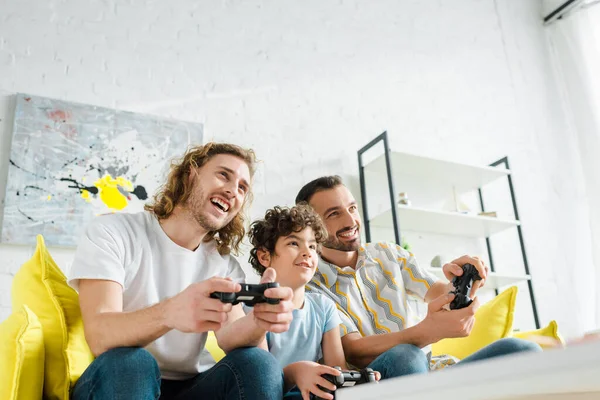 QUIIV, UCRÂNIA - JANEIRO 28, 2020: foco seletivo do casal homossexual feliz e do garoto de raça mista jogando videogame — Fotografia de Stock