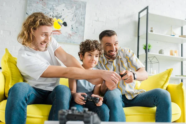 KYIV, UCRANIA - 28 de enero de 2020: enfoque selectivo de la feliz pareja homosexual jugando videojuegos con su hijo de raza mixta - foto de stock