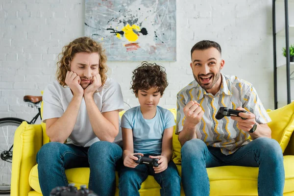 KYIV, UKRAINE - JANUARY 28, 2020: emotional homosexual couple playing video game with upset mixed race son — Stock Photo