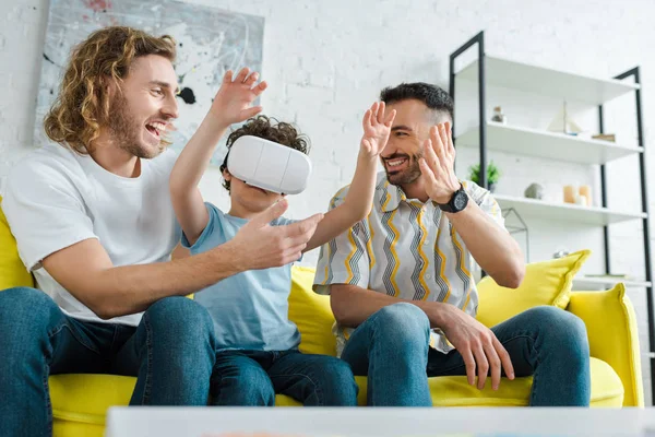 Feliz homosexual hombres gesto cerca mestizo hijo en virtual realidad auriculares - foto de stock