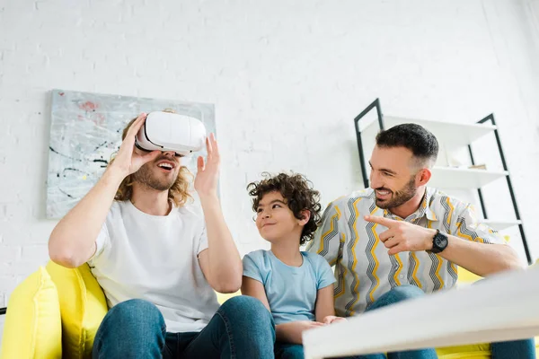 Enfoque selectivo de lindo hijo de raza mixta mirando al padre homosexual en auriculares de realidad virtual - foto de stock