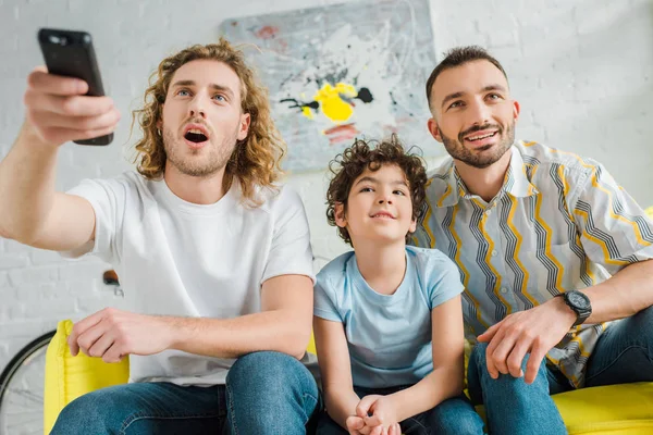 Selective focus of emotional homosexual parents and mixed race kid watching tv — Stock Photo