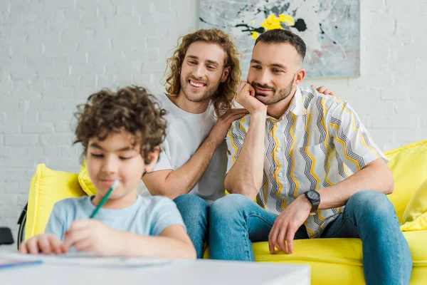 Selective focus of happy homosexual parents looking at mixed race son drawing at home — Stock Photo