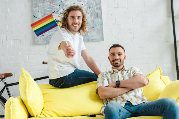 Веселые гомосексуальные мужчины, улыбающиеся возле флага lgbt в гостиной — стоковое фото