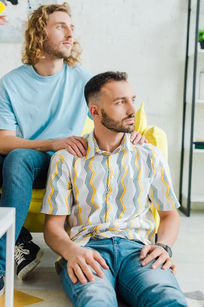 Hübsche homosexuelle Männer, die wegschauen, während sie im Wohnzimmer sitzen — Stockfoto