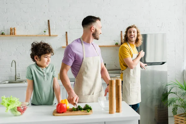Nettes Mischlingskind in der Nähe glücklicher homosexueller Eltern in der Küche — Stockfoto