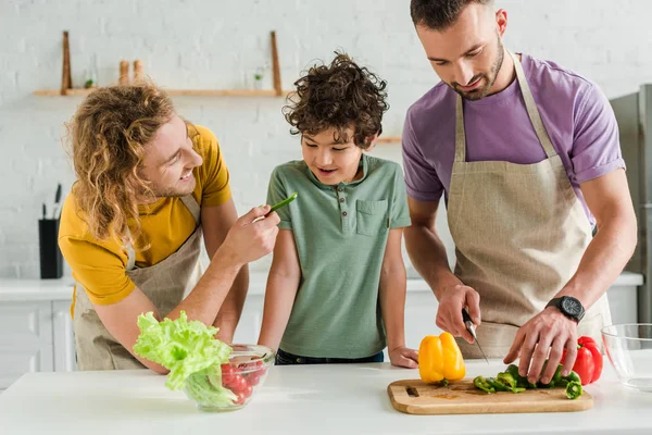 Carino ragazzo razza mista guardando padre omosessuale con peperone vicino partner cucina a casa — Foto stock