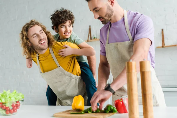 Feliz homosexual padre piggybacking mixta raza hijo cerca pareja cocinar en casa - foto de stock