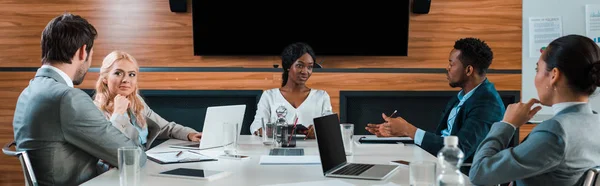 Panoramic shot of young multicultural businesspeople talking while sitting in conference hall — Stock Photo