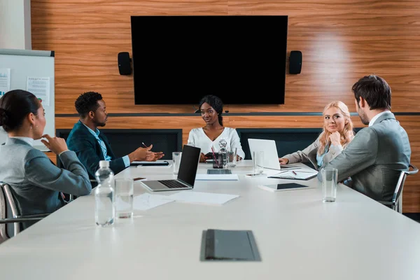Jóvenes empresarios multiculturales conversando durante el encuentro en sala de conferencias con pantalla LCD en la pared - foto de stock