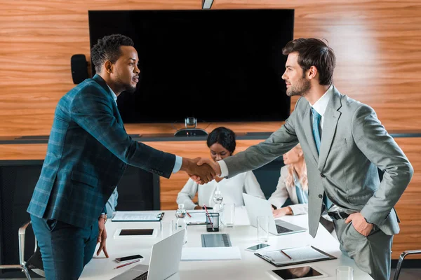 Dos hombres de negocios guapos dándose la mano mientras están de pie cerca de colegas multiculturales en la sala de conferencias - foto de stock