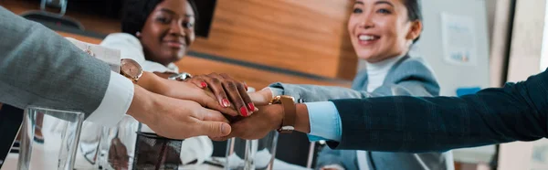 Vista cortada de jovens empresários multiculturais segurando as mãos unidas enquanto sentado na mesa na sala de conferências, tiro panorâmico — Fotografia de Stock