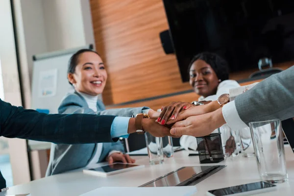 Enfoque selectivo de los jóvenes empresarios multiculturales tomados de la mano mientras están sentados en el escritorio en la sala de conferencias, vista recortada - foto de stock