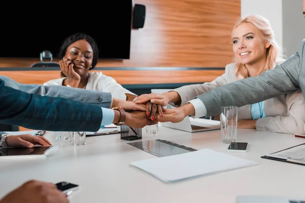 Cropped view of young multicultural businesspeople holding joined hands while sitting at desk in conference hall — Stock Photo