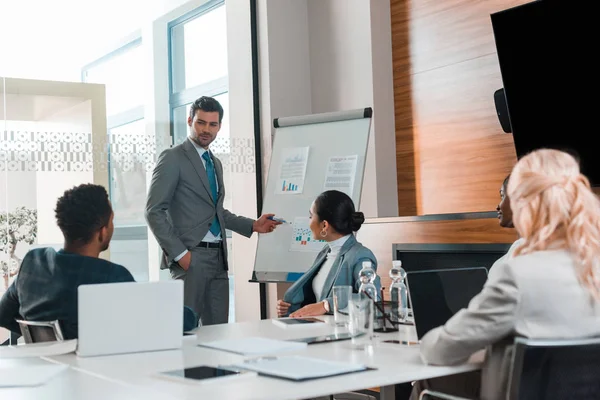 Empresários multiculturais olhando para o jovem empresário apontando com a mão para flipchart com infográficos — Fotografia de Stock