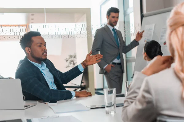 Bell'uomo d'affari che punta con mano a flipchart con infografica mentre colleghi multiculturali parlano in sala conferenze — Foto stock