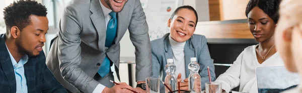 Обрізаний вид на молодого бізнесмена, що стоїть біля усміхнених мультикультурних колег, що сидять за столом у конференц-залі, панорамний знімок — стокове фото