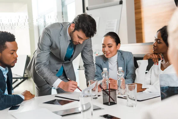 Selective focus of handsome businessman holding pencil while standing near multicultural colleagues sitting at desk in conference hall — Stock Photo