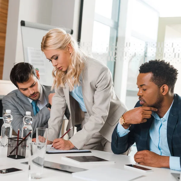 Attraktive Geschäftsfrau schreibt in Notizbuch neben aufmerksamen multikulturellen Kollegen, die am Schreibtisch im Konferenzsaal sitzen — Stockfoto