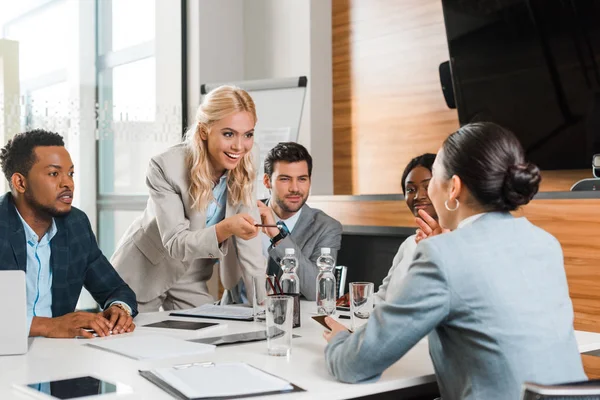 Young smiling businesswoman pointing with pencil near multicultural colleagues sitting at desk in conference hall — Stock Photo