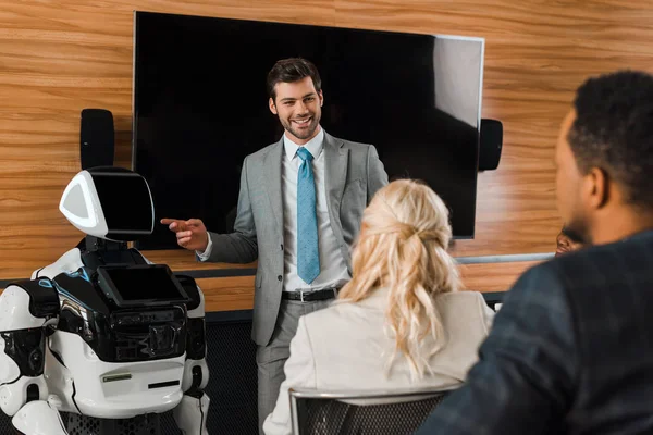 Uomo d'affari sorridente che punta il dito contro robot vicino a colleghi multiculturali — Foto stock