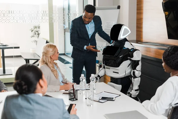 Belo empresário afro-americano apontando com a mão para robô perto de colegas multiculturais na sala de conferências — Fotografia de Stock