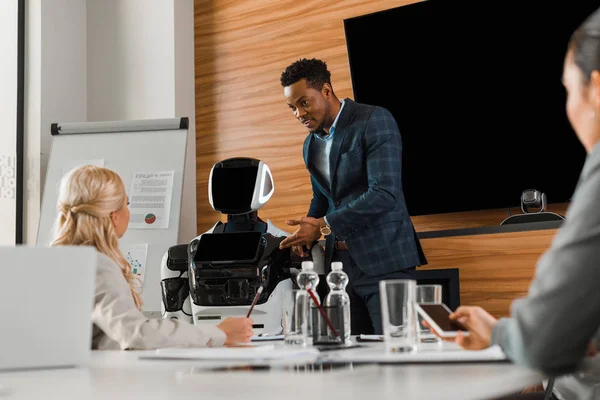 Красивый африканский бизнесмен указывает пальцем на робота рядом с коллегами в конференц-зале — стоковое фото