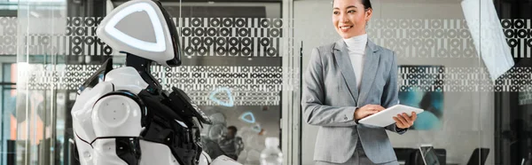 Panoramic shot of smiling asian businesswoman holding digital tablet while looking at robot — Stock Photo