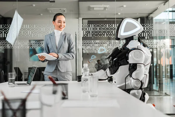 Selektiver Fokus einer lächelnden asiatischen Geschäftsfrau, die ein digitales Tablet in der Hand hält, während sie Roboter betrachtet — Stockfoto