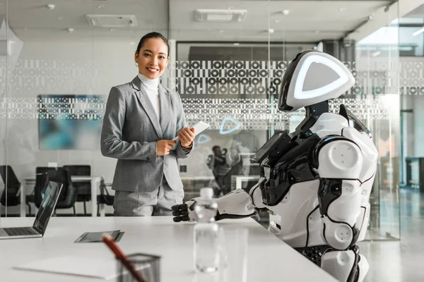Sonriente asiático mujer holding digital tablet mientras de pie cerca robot en sala de reuniones - foto de stock