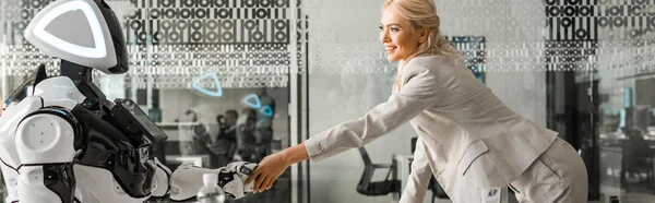 Panoramic shot smiling businesswoman shaking hands with robot sitting at desk in meeting room — Stock Photo