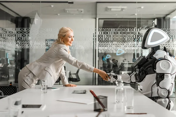 Усміхнена бізнес-леді тремтить руками з роботом, сидячи за столом в кімнаті для переговорів — стокове фото