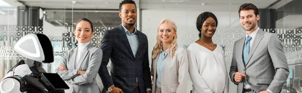 Panoramic shot of smiling multicultural businesspeople looking at camera while standing near robot — Stock Photo