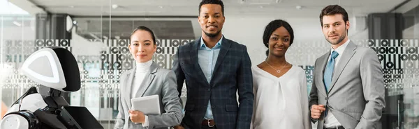 Panoramic shot of smiling multicultural businesspeople looking at camera while standing near robot — Stock Photo