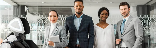 Tiro panorâmico de empresários multiculturais sorridentes olhando para a câmera enquanto estava perto do robô — Fotografia de Stock